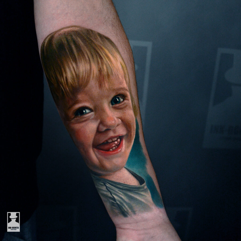 Kolorowy portret dziecka wykonany przez Marcina w realistycznym stylu. Autorski tatuaż w pełnym kolorze, z precyzyjnym odwzorowaniem emocji i detali, wykonany w INK-OGNITO Tattoo Rybnik.